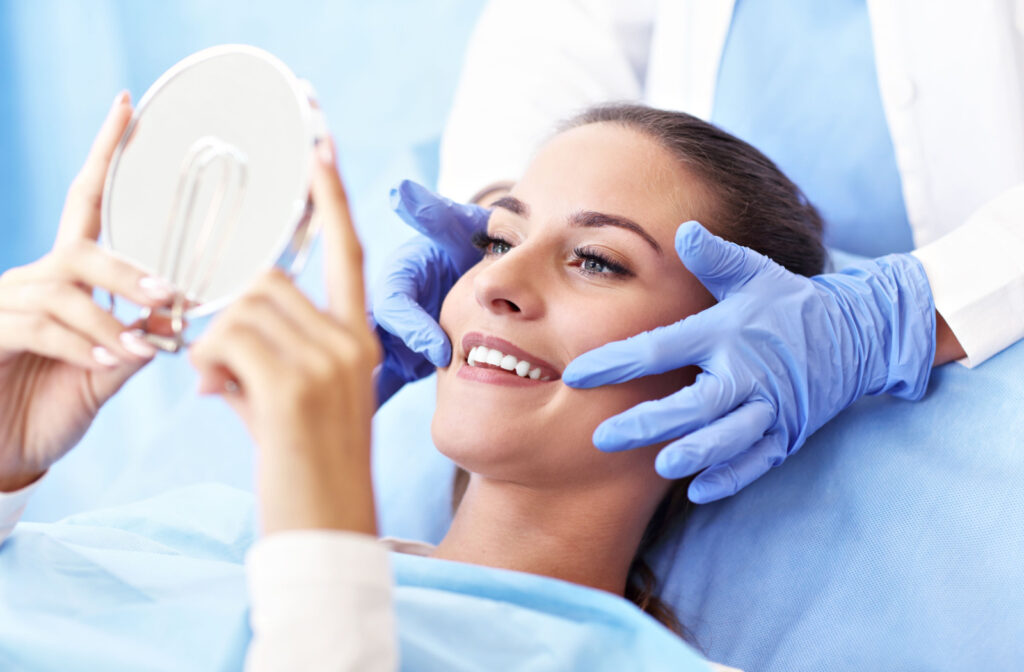 A dental patient looking in a handheld mirror, smiling after a professional dental cleaning as their dentists points to their mouth.